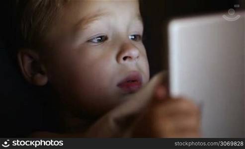 Close-up shot of little boy watching cartoon or movie on touch pad before bedtime