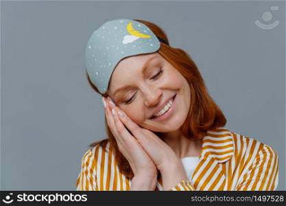 Close up shot of lovely tender woman has ginger hair, minimal makeup, leans at palms pressed together, smiles positively, wears blindfold on head, sees pleasant dreams. Healthy lifestyle concept