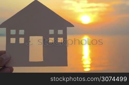 Close-up shot of male hand holding simple house model on background of golden sunset and sea with sun reflection. Villa or seaside house concept, copy space on the right