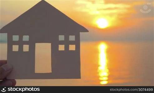 Close-up shot of male hand holding simple house model on background of golden sunset and sea with sun reflection. Villa or seaside house concept, copy space on the right