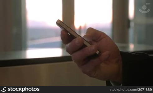 Close-up shot of male hand typing on smart phone at the airport. Blurred window with sunset view in background