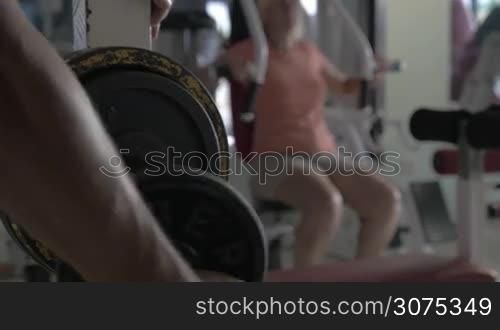 Close-up shot of man fixing extra weight disk on the bar-bell, woman training on exercise machine in background