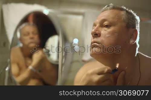 Close-up shot of mature man shaving in the bathroom with reflection in small round mirror