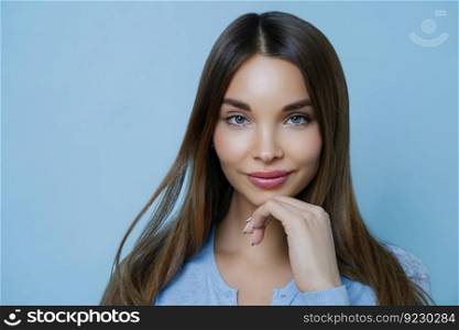 Close up shot of nice looking young European woman touches jawline gently, looks directly at camera, stands isolated over blue studio background, wears minimal makeup, admires something in front