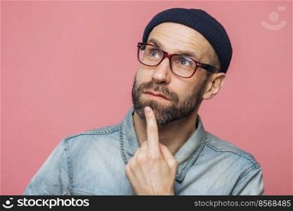 Close up shot of pleasant looking bearded male with thoughtful expression, keeps finger on chin, looks thoughtfully up, wears glasses and fashionable hat, poses against pink studio background