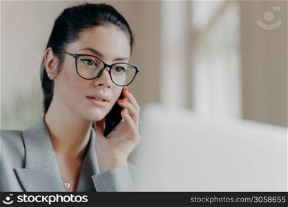 Close up shot of pleasant looking brunette European woman wears transparent glasses, focused in laptop screen, has telephone conversation during remote work, works on project. Technology concept