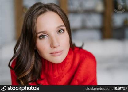 Close up shot of pleasant looking brunette young female has healthy skin, wears make up, knitted red sweater, poses against blurred background indoor with copy space for your promotional content
