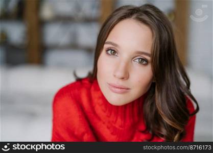 Close up shot of pleasant looking brunette young female has healthy skin, wears make up, knitted red sweater, poses against blurred background indoor with copy space for your promotional content