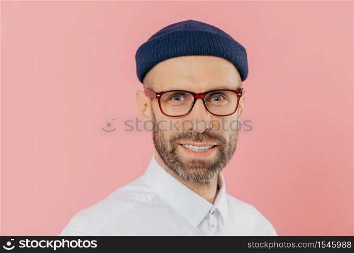 Close up shot of pleasant looking unshaven man wears glasses, has gentle smile, looks with satisfied facial expression at camera, wears white shirt, isolated over pink background. Male designer