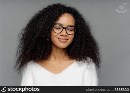 Close up shot of pleased smiling woman with bushy crisp hair, keeps eyes shut, smiles gently, wears optical glasses, imagines something pleasant, dressed in white jumper, isolated over dark grey wall