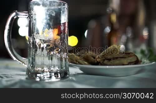 Close-up shot of pouring light beer with a lot of foam into glass mug next to plate with fried fish