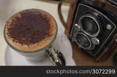 Close-up shot of pouring sugar into coffee and stirring up the foam, then taking foam with a spoon. Coffee cup standing near retro photo camera