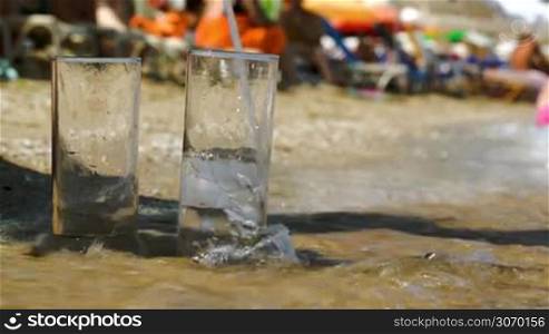 Close-up shot of pouring water into two glasses standing on the beach. Sea waves washing them. Refreshment on hot summer day