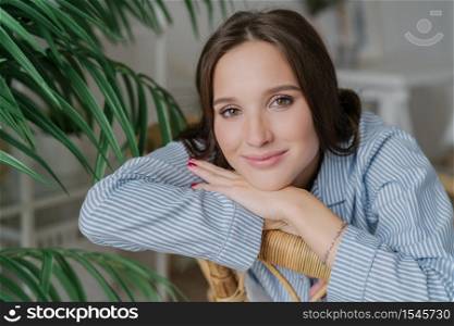 Close up shot of pretty dark haired young woman has charming gentle smile, leans on hands, poses indoor near green plantation, has red manicure, make up. People, leisure and relaxation concept