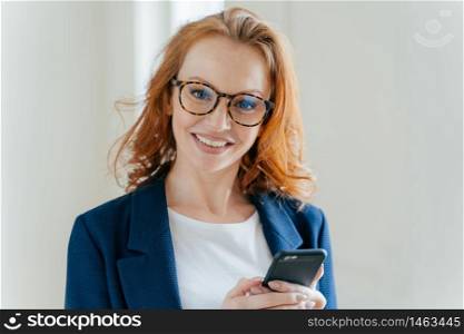 Close up shot of pretty redhead female directror has charming smile, dressed in elegant clothes, holds mobile phone, checks text message, surfs social networks, connected to wireless internet.