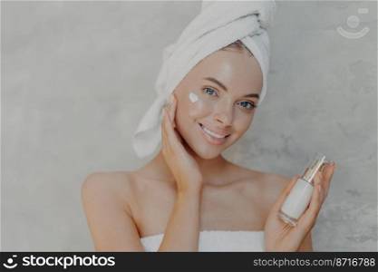 Close up shot of pretty young European woman with minimal makeup, applies moisturising cream on face, smiles gently and looks at camera, wears bath towel on head, isolated over grey background