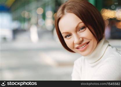 Close up shot of satisfied European woman with cheerful expression, dressed in casual clothes, looks mysteriously at camera, has outdoor walk, poses at street over blurred background with free space