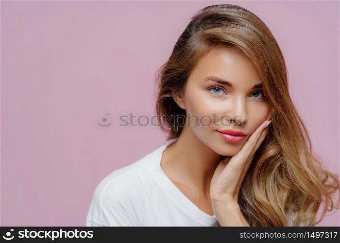 Close up shot of self confident beautiful Caucasian woman has long hair combed on one side, touches cheek with palm, wears minimal professional makeup, looks at herself in mirror, prepares for date