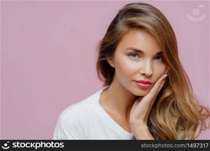 Close up shot of self confident beautiful Caucasian woman has long hair combed on one side, touches cheek with palm, wears minimal professional makeup, looks at herself in mirror, prepares for date