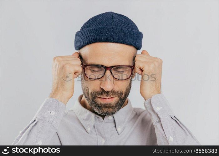 Close up shot of stressful unshaven man keeps hand near temples, has terrible headache, suffers from pain, wears headgear, spectacles, isolated over white background. Exhausted dissatisfied male