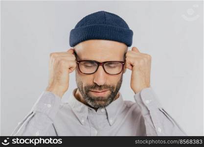 Close up shot of stressful unshaven man keeps hand near temples, has terrible headache, suffers from pain, wears headgear, spectacles, isolated over white background. Exhausted dissatisfied male