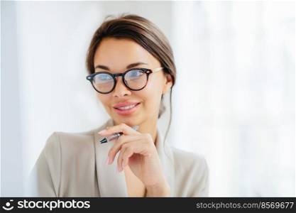 Close up shot of successful tender young woman with combed hair, keeps hand near chin, holds pen, focused in monitor, prepares business report, has manicure, dressed elegantly, works indoor.