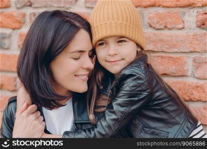 Close up shot of tender pleased young lady smiles gladfully and closes eyes, recieves hug from small daughter who wears leather jackt and hat, pose together over brick wall. Motherhood concept