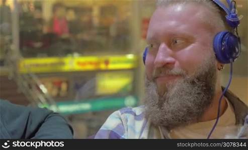 Close-up shot of two cheerful mature bearded men listening to music in headphone in shopping centre