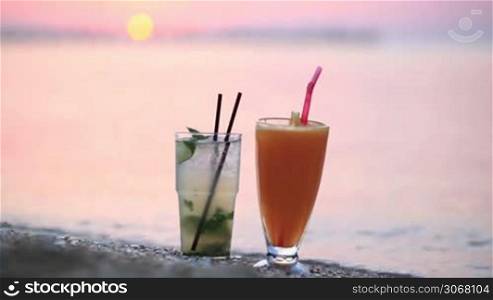 Close up shot of two fruity alcoholic cocktails mojito and orange juice standing side by side in their tall glasses in the sand on a tropical beach at sunset against a colourful pink sky in the evening