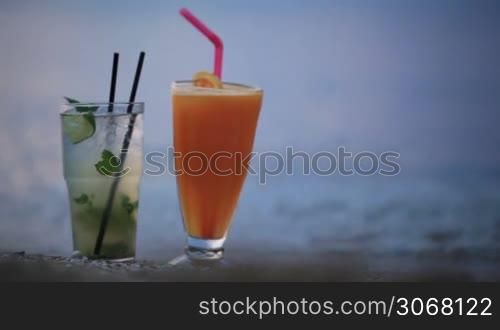 Close up shot of two fruity alcoholic cocktails mojito and orange juice standing side by side in their tall glasses in the sand on a tropical beach at the evening with copy space from right