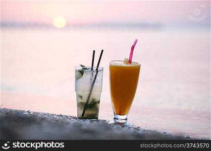 Close up shot of two fruity alcoholic cocktails mojito and orange juice standing side by side in their tall glasses in the sand on a tropical beach at sunset against a colourful pink sky in the eveningcktails at sunset