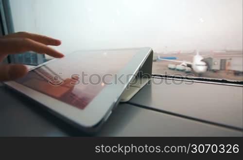 Close-up shot of woman hand typing on tablet PC by the window. Airport area and boarding plane in background