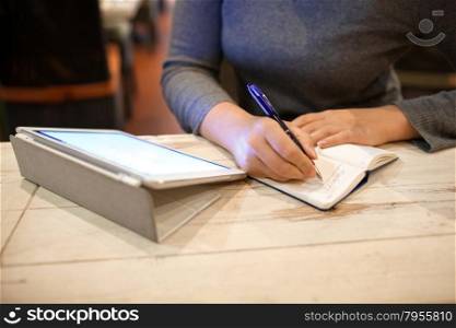 Close-up shot of woman in cafe putting down some information found by means of tablet computer. Woman taking down information in notebook