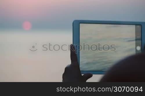 Close-up shot of woman taking pictures of sunset over sea using tabler computer. At first focus on pad, then on sea and sunset