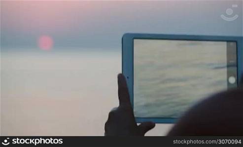 Close-up shot of woman taking pictures of sunset over sea using tabler computer. At first focus on pad, then on sea and sunset