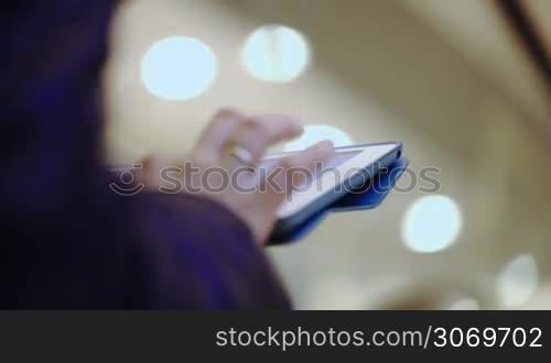 Close-up shot of woman typing on her tablet while riding on escalator. Defocused lights in background