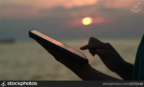 Close-up shot of woman using tablet computer outdoor. Sunset over the sea in background