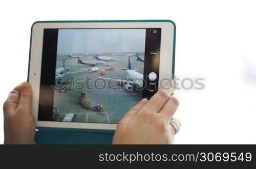 Close-up shot of woman using touch pad for taking pictures of airport area with planes and trucks through the window