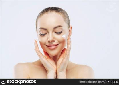 Close up shot of young adult woman with naked shoulders applies nourishing moisturising cream for soft pure skin, has tender expression, isolated on white background. Beauty, cosmetology concept