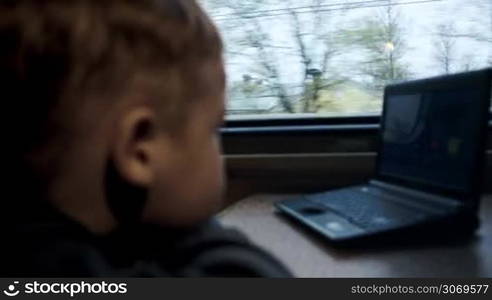 Close-up side view of a boy watching movie or cartoon on laptop during his traveling in the train. Focus on the window with passing trees and houses