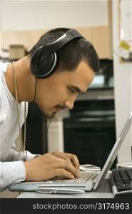Close-up side view of Asian young adult man leaning over laptop typing on keyboard wearing headphones.