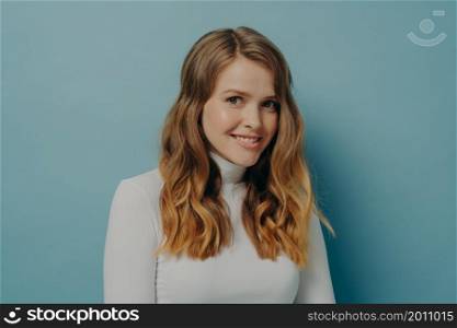 Close-up sideways photo of young charming lady in white turtleneck with wavy dyed hair looking at camera smiling brightly with head slightly tilted isolated over pastel blue studio background. Young charming happy woman looking at camera smiling brightly and expressing positive emotions