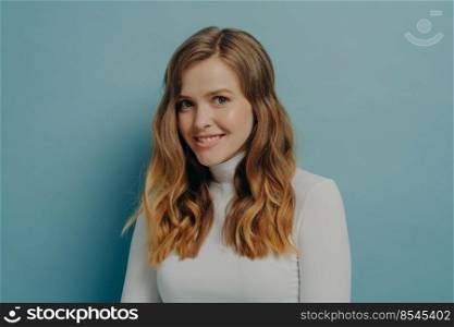 Close-up sideways photo of young charming lady in white turtleneck with wavy dyed hair looking at camera smiling brightly with head slightly tilted isolated over pastel blue studio background. Young charming happy woman looking at camera smiling brightly and expressing positive emotions