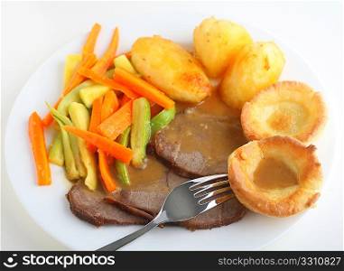 Close-up, slanted view of slices of roast beef with yorkshire pudding, roasted potatoes and sauteed carrots and courgettes, topped with a rich gravy