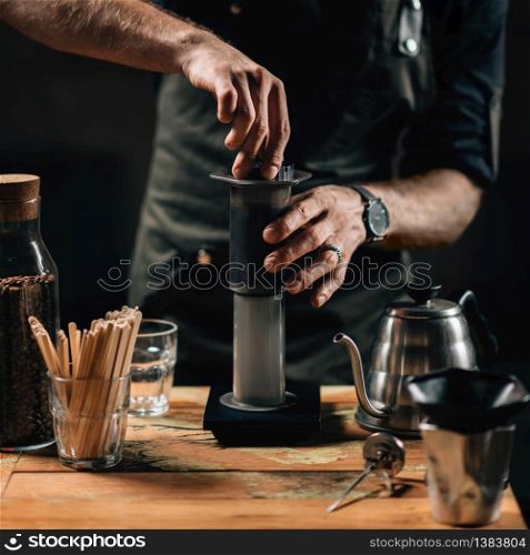 Close up square image of barista making air press coffee. Barista with tattooed arms wearing dark uniform.
