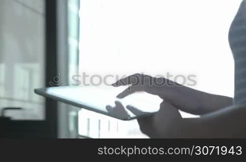 Close-up steadicam shot of a walking girl typing on tablet computer in sunlight coming through the big windows