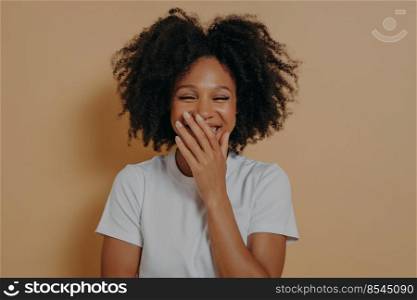 Close up studio portrait of happy biracial girl. Beautiful dark skinned millennial woman covering her mouth with hand while laughing. Cute positive mixed race female isolated over beige background. Beautiful dark skinned millennial woman covering her mouth with hand while laughing