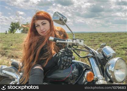 close up The red-haired biker girl is sitting on a motorcycle. field of meadow and clouds
