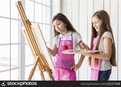 close up two girls standing near window painting easel with paint brush