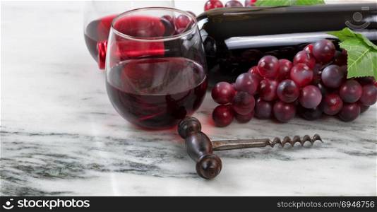 Close up view of a corkscrew with red wine bottle, filled glasses and grapes on natural marble stone setting in background